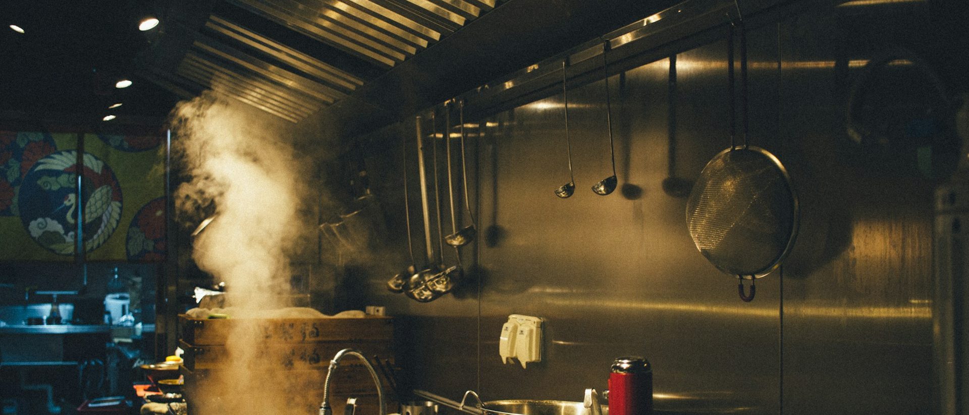 Steam rising from bamboo steamers in a working kitchen, ladles and a strainer hanging along the pass.